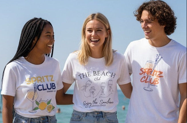 Three people standing outdoors by the ocean wearing white graphic T-shirts with cocktail-themed prints and text including 'SPRITZ CLUB' and 'THE BEACH Club'.